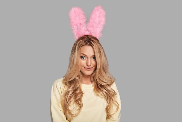 Playful beauty. Attractive young woman in pink bunny ears looking at camera and smiling while standing against grey background