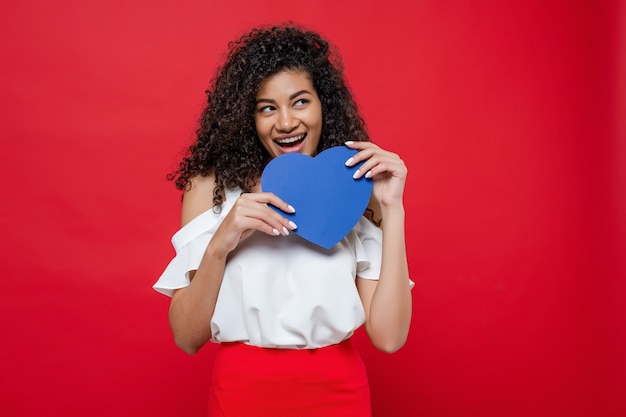 Playful black woman with blue heart shape in hands isolated over red