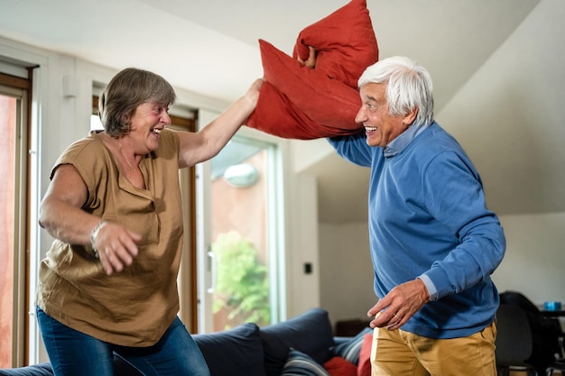 Playful and carefree senior couple making pillows fight in the middle of living room leisure lifestyle and happiness concept in silver age