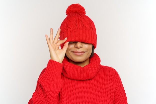 Playful funny african woman has fun wearing winter hat and sweater cover eyes under cap studio shot