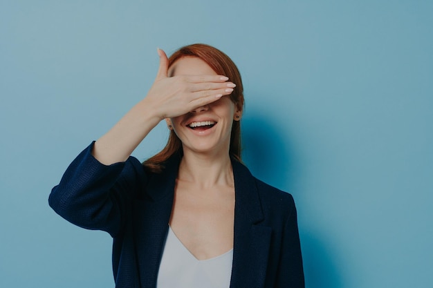 Playful funny ginger girl covering her eyes with palm isolated on blue background isolated on blue