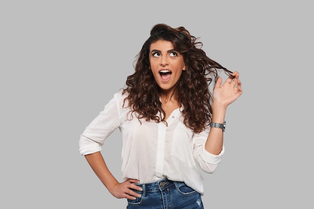 Playful girl. Attractive young woman playing with her hair and keeping mouth open while standing against grey background