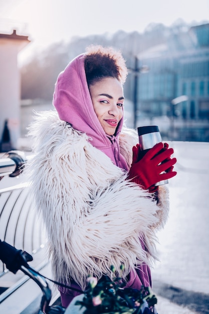 Playful glance. Charming female keeping smile on her face while holding cup in both hands