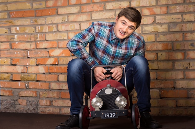 Playful handsome young man having fun riding a vintage toy truck in front of a wall looking at the camera with a happy smile