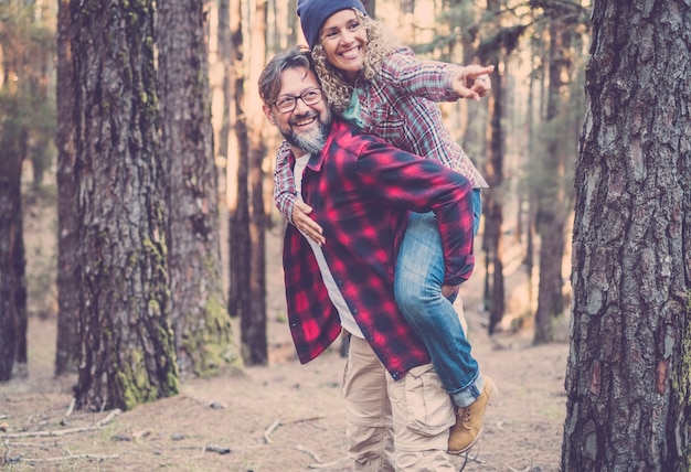 Playful happy handsome caucasian couple having piggyback while walking on trail in woods. Adventure in nature concept - young adult man and woman enjoy together outdoor leisure