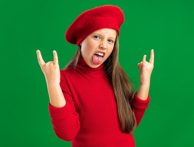 Playful little blonde girl wearing red beret doing rock sign showing tongue looking at camera isolated on green wall