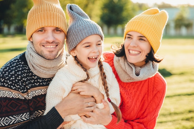 Playful little child with pigtails wears warm clothes spends free time with lovely affectionate parents have happy expression feel relaxed Three young family members enjoy pleasant moments