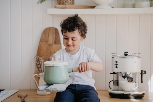 Playful little curly boy in white tshirt and blue pants sits on kitchen desk with potholder on hand