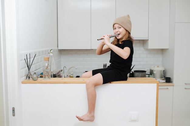 Bambina giocosa con un cappello beige che gioca con un flauto, seduta su un tavolo alto in cucina a casa.
