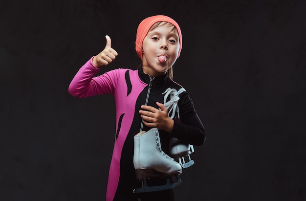 Playful little girl dressed in sportswear holds ice skates shows the pulled out tongue and thumbs up. Isolated on a dark textured background.