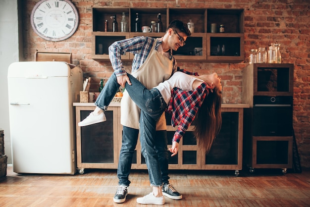 遊び心のある愛のカップルは、キッチンでポーズします。エプロンと若い女性の男が踊って、面白い料理