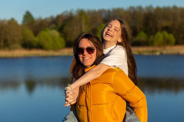 Playful mother giving daughter piggy back ride at spring lake shore