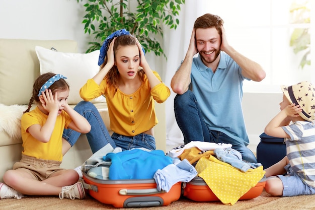 Playful parents with little kids packing luggage sitting on\
floor and covering ears in amazement looking unready for trip