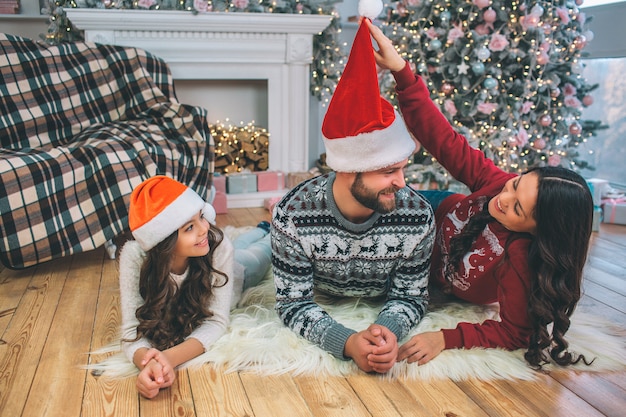 Playful and positive young woman holds end on red hat and looks at her husband. He is lying on floor and smiles. Guy look at wife. Their daughter lying besides father.