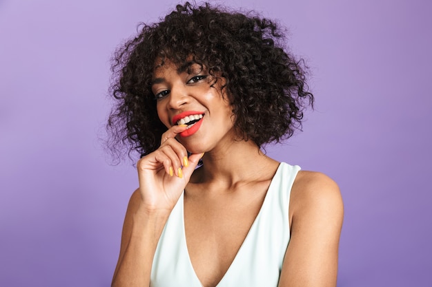 Playful pretty african woman bites her finger and looking directly over violet background