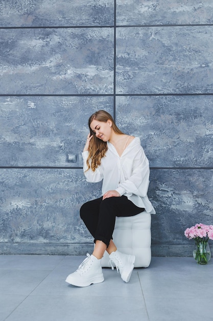 Playful selfconfident woman in a stylish cotton white shirt black jeans white leather sneakers casual styleBlondhaired woman on a gray background in stylish clothes