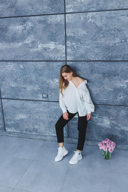 Playful selfconfident woman in a stylish cotton white shirt black jeans white leather sneakers casual styleBlondhaired woman on a gray background in stylish clothes