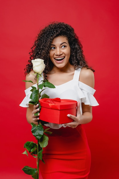 Playful smiling black woman holding white rose and heart shaped gift box isolated on red