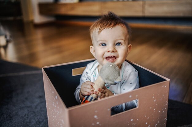 Игривый улыбающийся кавказский очаровательный маленький белокурый мальчик сидит в коробке и играет со своей любимой игрушкой. Домашний интерьер.