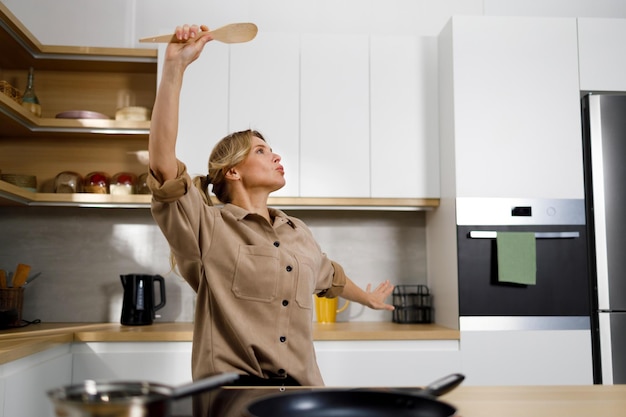 Foto donna giocosa che balla con la spatola di legno mentre cucina nella cucina di casa