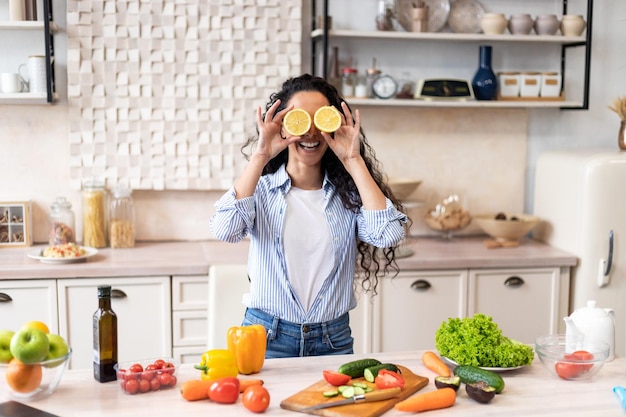 Playful woman having fun putting orange slices to eyes at table with colorful vegetables in kitchen