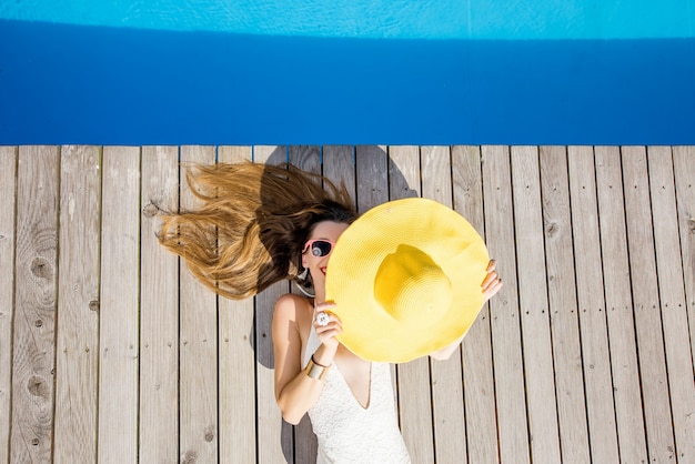 プールサイドで黄色い日よけ帽で横になっている遊び心のある女性。夏休みのコンセプト