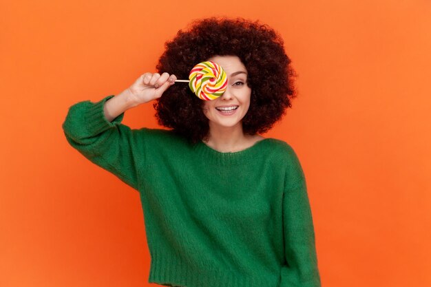 Playful woman with Afro hairstyle wearing green casual style sweater covering mouth with multicolor lollipop having fun childish behavior Indoor studio shot isolated on orange background