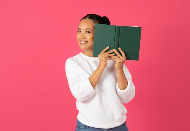 Photo playful young asian woman holding book in front of her face