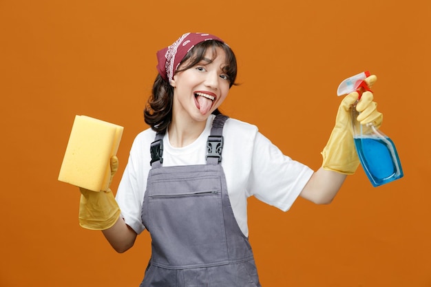 Playful young female cleaner wearing uniform rubber gloves and bandana holding sponge and cleanser looking at camera showing tongue isolated on orange background