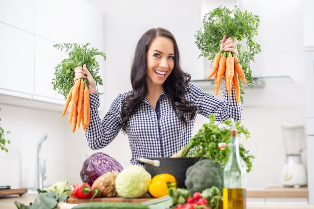 Playful young woman in her kitchen holding fresh carrot in both hands - diet vegetable and heath concept.