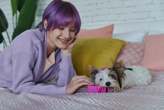 Игривая молодая женщина держит маленькую подарочную коробку, лежа в постели со своей собакой