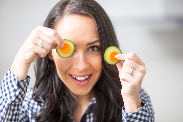 Playful young woman holding slices of carrot with cucumber in the kitchen - diet vegetable and heath concept.