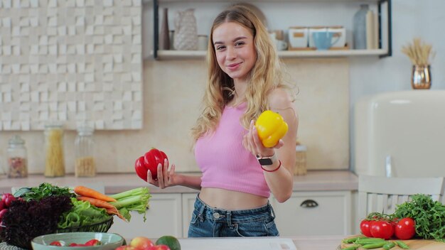 Playful young woman smiling covering eyes by yellow and red pepper in the home kitchen Healthy lifestyle and dieting concept