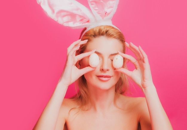 Playful young woman wearing bunny ears and holding up a colorful easter egg in front of her eye