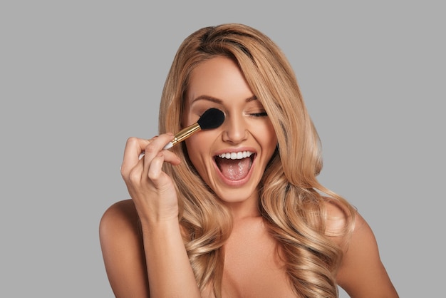 Playful young woman with fresh glowing skin applying beauty product and smiling while standing against grey background