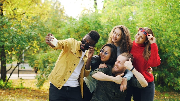 Playful youth men and women are taking selfie in park using smartphone making funny faces and wearing sunglasses standing outdoors and laughing Fun and nature concept