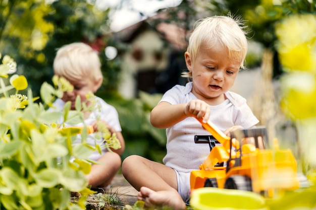 プラスチック製の掘削機ゲームで庭で遊ぶ。幼児の双子は庭に座って植物で遊んでいます。ブロンドの髪と大きな青い目を持つ子供は、お気に入りのおもちゃで遊んでいるように見えます