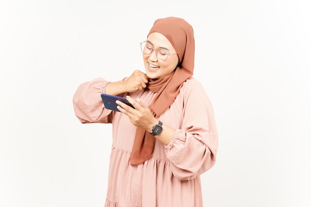 白で隔離されるヒジャブを身に着けている美しいアジアの女性のスマート フォンでモバイル ゲームをプレイ