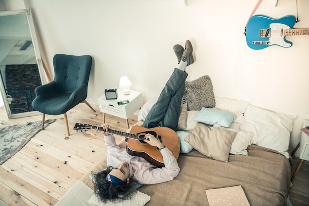 Photo playing sad songs. dark-haired stylish musician chilling upside down on covered bed and performing sounds on musical instrument