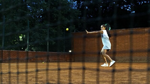 Playing tennis at night young girl blocking the ball with the tennis racket during the training