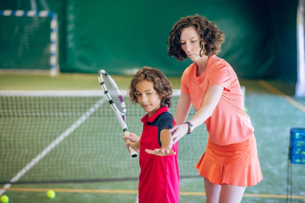 Playing tennis. Woman in bright clothes teaching a boy how to hit a tennis ball