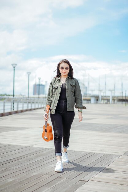 Playing Ukulele of Young Beautiful Asian Woman Wearing Jacket And Black Jeans Posing Outdoors