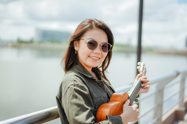 Playing Ukulele of Young Beautiful Asian Woman Wearing Jacket And Black Jeans Posing Outdoors