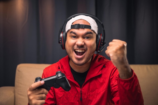 Playing video game Young asian handsome man sitting on sofa holding joystick in living room Happiness Streamer Indian man wearing headset playing game online in the darkroom