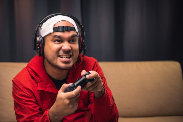 Playing video game young asian handsome man sitting on sofa\
holding joystick in living room happiness streamer indian man\
wearing headset playing game online in the darkroom