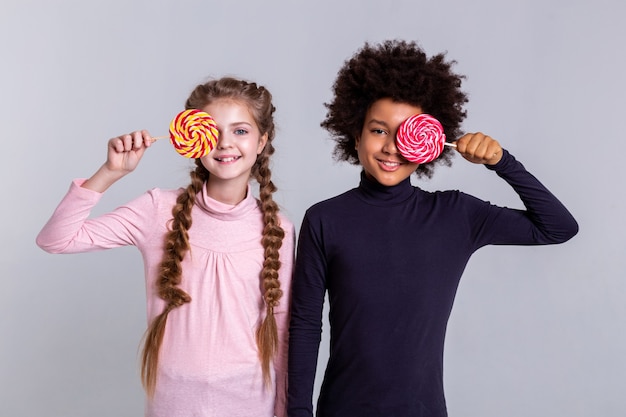 Playing with candies. Smiling beaming kids wearing turtlenecks while making photos and holding colorful candies in front of faces