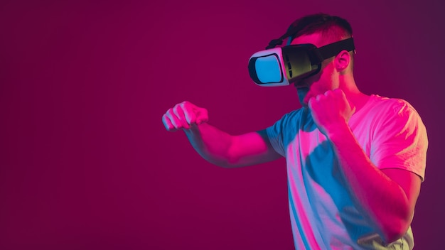 Playing with VR, shoting, driving. Caucasian man's portrait isolated on pink-purple  wall in neon light. Male model with devices. Concept of human emotions, facial expression,