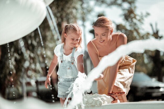 Playing with water. Beautiful smiling woman playing with her little happy daughter near the fountain.