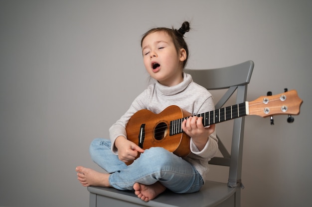 Plays the ukulele and sings. Early childhood development. Beautiful little girl practicing singing and playing the guitar