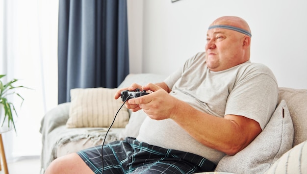 Plays video game Funny overweight man in casual clothes is indoors at home